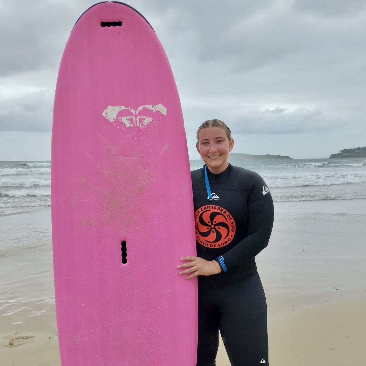 A student posing with a pink surf board at the beach in Spain