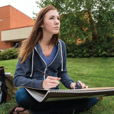 Student sitting on grass drawing with a pad and pencil