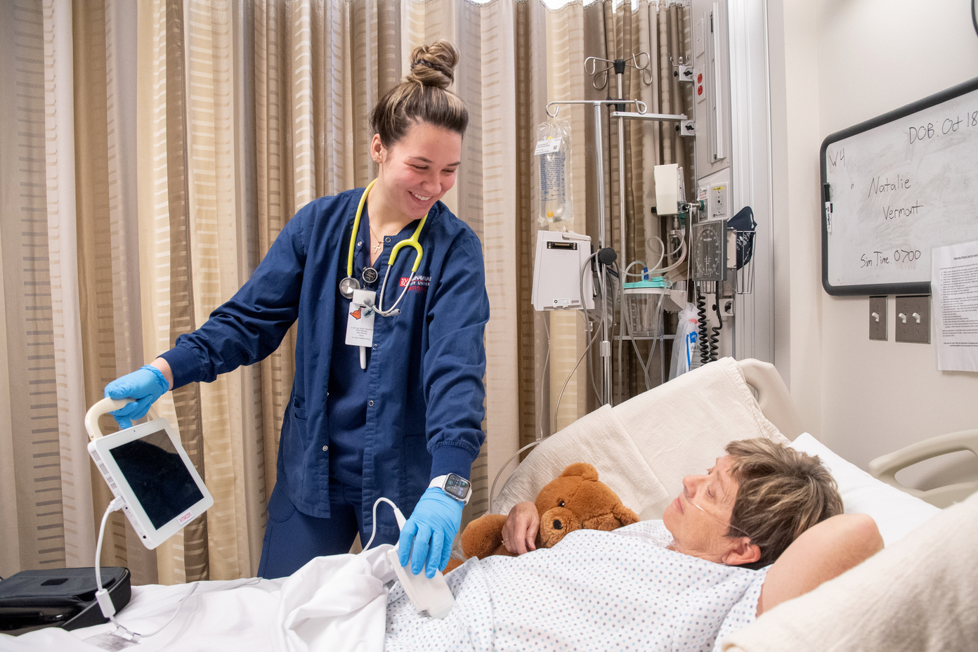 Young woman in scrubs stands next to a hospital patient's bedside